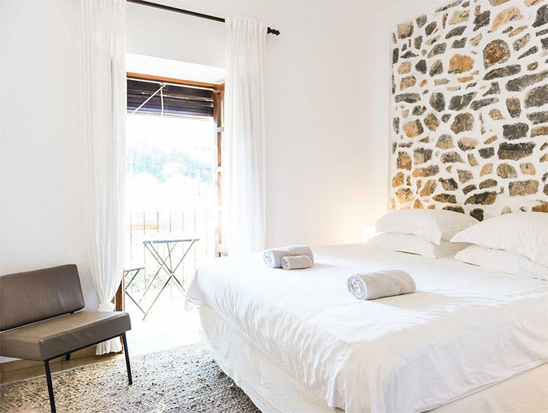 Bright bedroom with white bedding, rolled towels, stone accent wall, and balcony with outdoor table