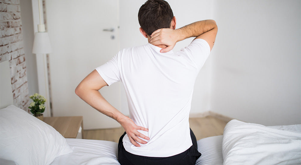 Man in white t-shirt sitting on bed holding neck and lower back in pain in bright bedroom